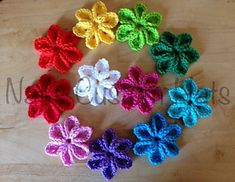 six crocheted flowers arranged in a circle on a wooden surface, with the petals colored red, yellow, green, blue, and white