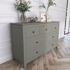 a dresser with flowers and pictures on it in a white walled room next to a wooden floor