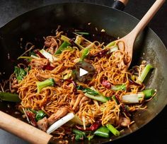 a wok filled with noodles and vegetables on top of a table next to two wooden spoons