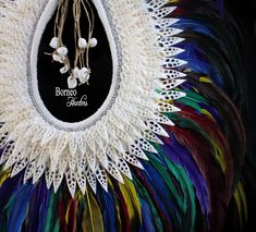 A handmade Papua New Guinea tribal inspired large white braided Rope necklace, decorated with one hundred and forty seven white Shell slices, a band of round silver beads, and a fringe of purple, yellow, green, blue, red and iridescent black dyed Feathers. The inner width of the band of the necklace is 5.25 inches, and the maximum outer width of the necklace is 23 inches. The drop length from the top of the necklace to the bottom of the feathers,is 27 inches. The white Shells are, between 1.3 an White Bohemian Woven Jewelry, Bohemian White Woven Jewelry, Handmade White Bohemian Necklaces, White Handwoven Festival Necklace, White Handwoven Necklace For Festivals, White Artisan Beaded Necklaces For Festivals, Artisan White Beaded Necklaces For Festivals, White Artisan Beaded Necklace For Festivals, Artisan White Beaded Necklace For Festivals