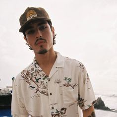 a man in a hawaiian shirt and hat standing by the ocean with his hand on his hip