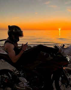 a man sitting on a motorcycle next to the ocean at sunset with his cell phone in his hand