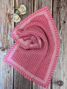 a pink crocheted blanket sitting on top of a wooden table next to a white flower