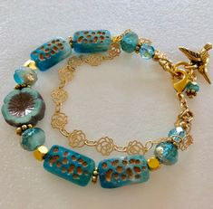 Lovely double strand beaded Premium Czech glass flower bracelet featuring table cut matte aqua with bronze rectangular beads, gold etched ivory/aqua rondelles, fire polished aqua Aurora Borealis crystals, and single 14 mm turquoise Picasso pansy flower. All accented with antique gold plated daisies and shiny gold cubes. Satin Hamilton rose chain adds the perfect delicate second strand. Premium gold electroplated brass chain in satin finish with intricate rose flower pattern. Bracelet is beaded o Hand-strung Gold Czech Glass Jewelry, Hand-strung Gold Jewelry With Czech Glass, Gold Czech Glass Jewelry, Hand-strung, Gold Hand-strung Czech Glass Bracelets, Gold Czech Glass Hand-strung Bracelet, Unique Gold Beaded Bracelets Nickel-free, Unique Gold Beaded Bracelets Nickel Free, Blue Double Strand Bracelets As Gift, Paris Charm Bracelet