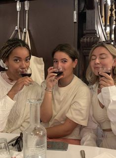 three women sitting at a table drinking from wine glasses in front of their faces and smiling