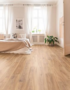 a bedroom with wood flooring and white curtains on the window sill, next to a bed