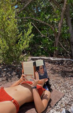 a woman laying on top of a towel reading a book next to trees and bushes