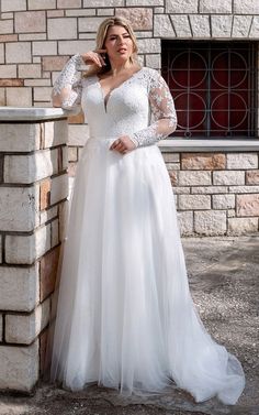 a woman standing next to a brick wall wearing a white wedding dress with long sleeves