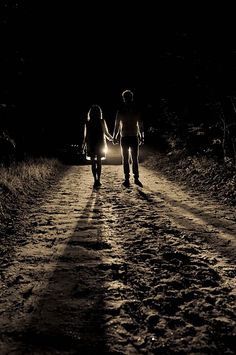 two people walking down a dirt road at night with the headlights on and one person holding hands