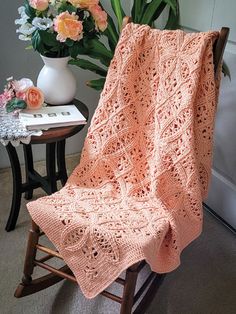 a pink crocheted blanket sitting on top of a rocking chair next to a vase with flowers