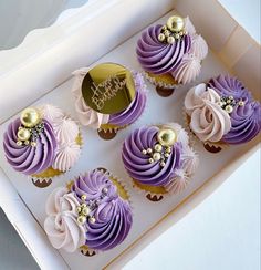 cupcakes with purple frosting and gold decorations in a white box on a table