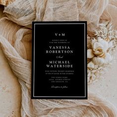 an elegant black and white wedding card on top of a cloth with flowers in the background