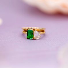 an emerald and diamond ring sitting on top of a purple surface with flowers in the background