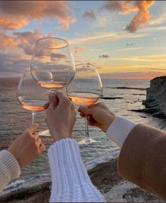 two people toasting wine glasses with the ocean and sky in the background at sunset