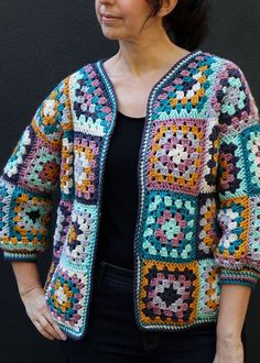 a woman standing in front of a black wall wearing a colorful crocheted jacket