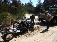 two men standing next to an off road vehicle in the sand with another man looking at it