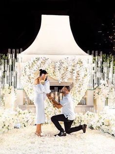 a man kneeling down next to a woman in front of a white flower covered wall