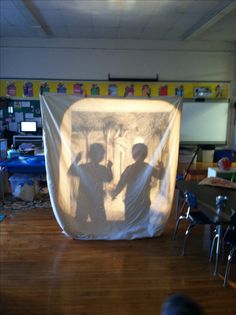 the shadow of two children in front of a white screen on a wooden floor with chairs