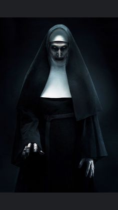 a nun with white makeup and black robes is standing in the dark, holding her hands out