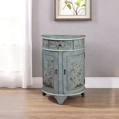 a small blue cabinet with flowers painted on the doors and drawers is in front of a window