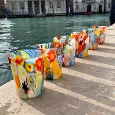 several colorful cups are lined up on the edge of a pier near water with buildings in the background