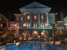 people are standing in front of a large house at night with the pool lit up