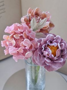 two pink flowers in a glass vase on a table with a book behind the vase