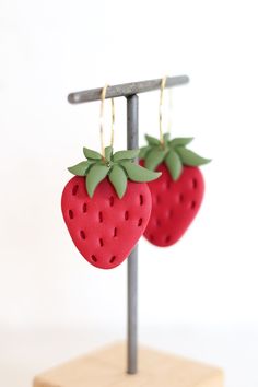 two red strawberries are hanging from a metal stand on a white background, one is shaped like a strawberry and the other has green leaves