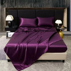 a bed with purple sheets and pillows in a room next to a night stand or lamp