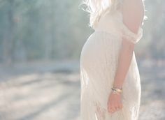 a pregnant woman in a white dress is holding her hand up to her head while standing on a dirt road