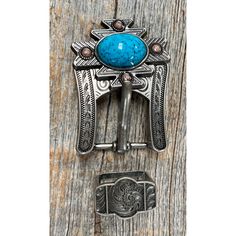 a belt buckle with a turquoise stone in the center on a wooden surface, next to a metal clip