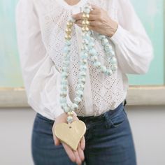 a woman wearing a white blouse and blue jeans holding a gold heart necklace in her hands