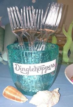 there is a blue glass cup with silver forks in it and some seashells on the table