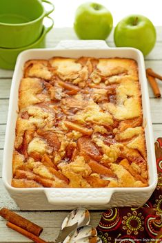 a casserole dish with apples and cinnamon on the side next to two green mugs