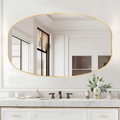 a white bathroom with gold trim and marble counter tops, an oval mirror above the sink