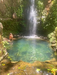 a person sitting in the water next to a waterfall with a man standing on it