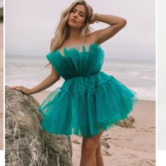 two women in dresses standing on the beach and one is leaning against a rock with her hands behind her head