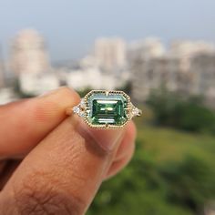 a person holding an emerald and diamond ring in front of a cityscape background