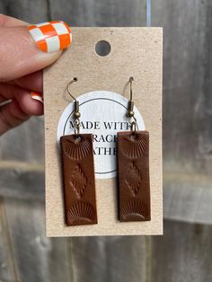 a pair of brown leather earrings is shown in front of a wooden card with an orange and white striped background