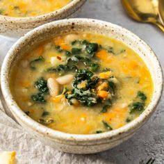 two bowls of soup with spinach, carrots and cheese on the table next to gold spoons