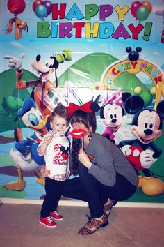 a woman holding a small child in front of a mickey mouse birthday backdrop with balloons