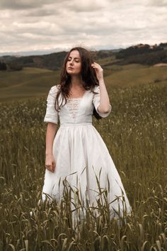 a woman in a white dress is standing in a field with tall grass and looking off to the side