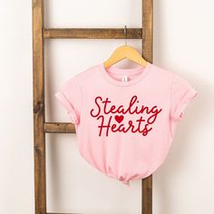 Looking for a cute tee for your kids? We have the perfect Stealing Hearts Cursive graphic tee addition to their closet! Also available in youth tees. Valentine's Day Graphic Tee With Slogan, Cute Cotton T-shirt With Heart Shape, Cute Cotton T-shirt With Heart Design, Cute Heart-shaped Cotton T-shirt, Cute Slogan Tops For Valentine's Day, Cute Cotton T-shirt With Heart Graphic, Valentine's Day Cute Slogan Tops, Valentine's Day Graphic Cotton T-shirt, Family Matching Graphic Print Tops For Valentine's Day