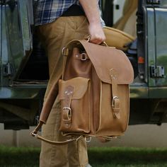 A man with a squared leather backpack  in tobacco leather in his hand, wearing bright pants in front of a car Classic Rectangular Laptop Bag For Outdoors, Classic Rectangular Laptop Bag For Outdoor, Classic Rectangular Outdoor Laptop Bag, Standard Backpack With Adjustable Strap For Adventure, Classic Rectangular Briefcase For Outdoor, Rectangular Leather Backpack For Outdoor Activities, Rectangular Adventure Bag, Classic Leather Backpack For Adventure, Classic Leather Adventure Backpack