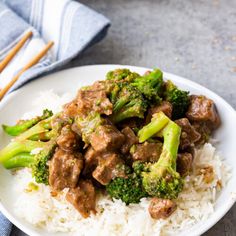 a white plate topped with beef and broccoli on top of rice next to chopsticks