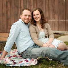 a man and woman sitting on a blanket in front of a wooden fence smiling at the camera