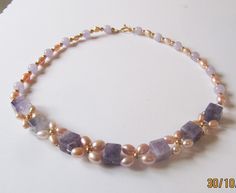 Pearl and Amethyst necklace woven into a floral pattern. Large cube of Amethyst has been woven with graduated sized rice Cultured Freshwater pearls in a lovely Apricot/Peach colour. The largest ones are at the front and measure 6x9mm to 7/8x5mm. The overall effect is a vintage style necklace with high visual impact.The cubes of Amethyst are substantial and measure 10mm. The other Amethyst are rounds and measure 8mm. They are matched with rice pearls measuring graduated from 9x6mm to 8x5mm. All t Purple Pearl Jewelry With Gemstone Beads, Handmade Elegant Amethyst Beaded Necklaces, Purple Pearl Jewelry With Natural Stones, Handmade Delicate Purple Necklace, Elegant Lavender Necklaces With Gemstone Beads, Wedding Necklaces With Amethyst And Natural Stones, Amethyst Necklaces With Natural Stones For Weddings, Purple Single Strand Pearl Jewelry, Elegant Handmade Lavender Necklaces