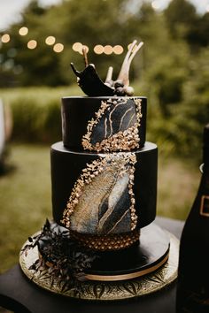 a three tiered cake with black and gold designs on it sitting on a table next to a bottle of wine