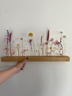 a person is holding a wooden shelf with flowers on it and one hand is pointing at the wall