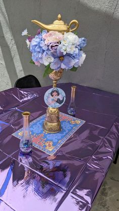 a purple table topped with a vase filled with blue flowers and a golden teapot